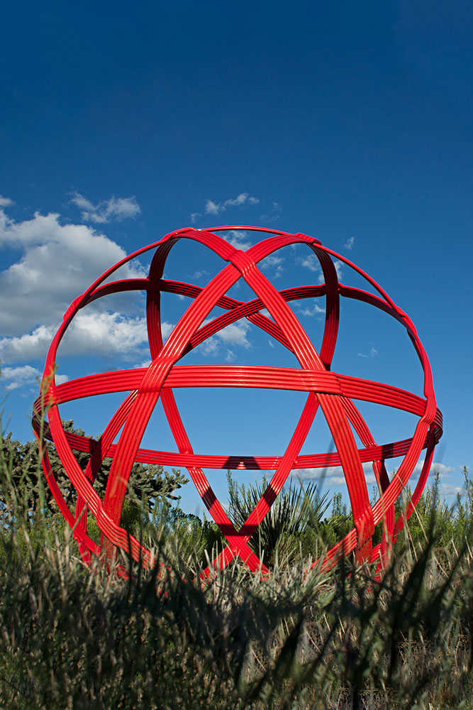A 51-inch red steel orb sculpture representing a wicker ball, displayed in a field of grass. Hand-forged by New Mexico blacksmith Christopher Thomson.