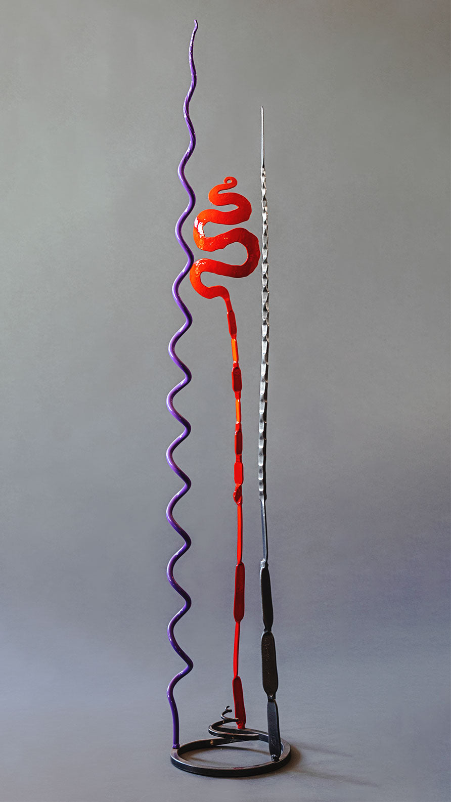 Abstract metal sculpture with three slender pieces affixed to a spiral base. The three pieces are in the colors lavender, red and metallic black
