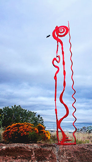 A red outdoor garden sculpture with abstract shapes. A hummingbird is hovering nearby.