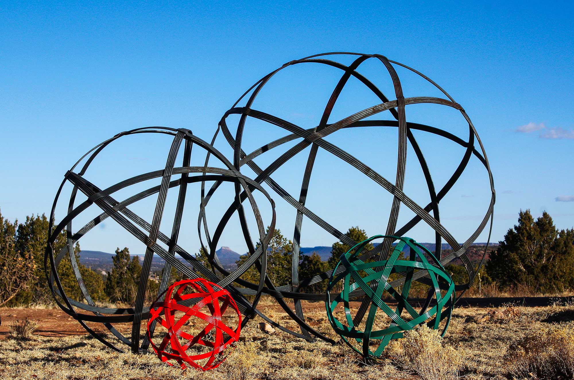 Large hand-forged steel orb sculptures resembling chinlone wicker balls, called "Chinlone Garden Orb Sculptures," displayed on a high desert landscape. The sculptures come in various sizes.