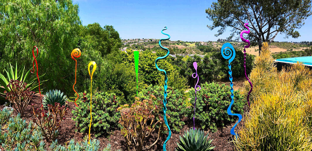 Eight colorful metal garden sculptures by Christopher Thomson decorate a garden scene.