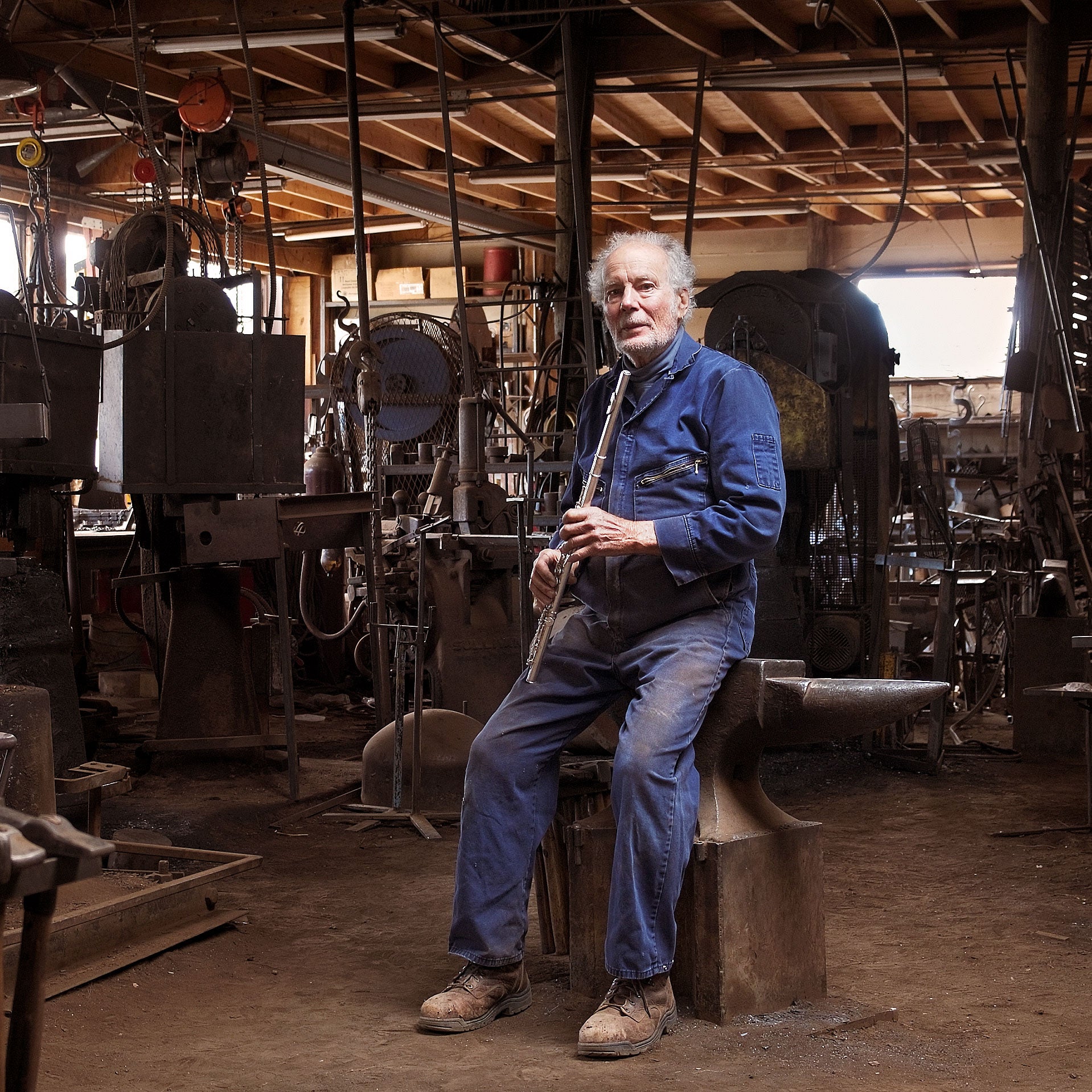Christopher Thomson sitting on an anvil while holding a flute in his blacksmith workshop filled with machinery and tools