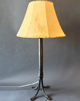 Hand-forged Collard Sheave wrought iron lamp featuring a tapered, black iron base with four elegantly curved feet. The lamp has a warm, tan lampshade with stitched edges, contrasting the dark metal and giving it a rustic yet refined appearance.