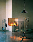 A modern fireplace with a lit fire, set in a minimalist interior with a standing fireplace tool set, glass-top table, hanging pendant light, and decorative metal sculptures, all hand forged by blacksmith, Christopher Thomson Ironworks.