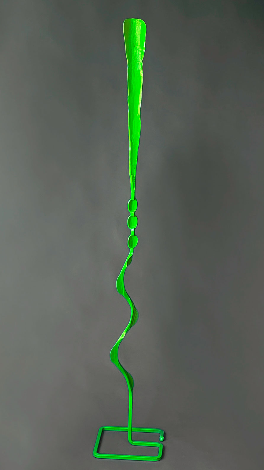 A bright green single element abstract sculpture made of hand forged steel. The sculpture has a square base and stands alone.