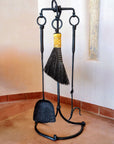 A rustic black iron fireplace tool set with a stand, including a shovel, poker, and broom displayed in a corner with terracotta tile flooring and cream walls.