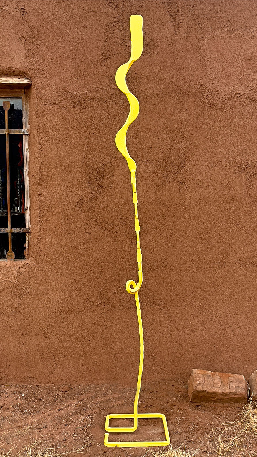 A pineapple yellow free standing sculpture, minimal in design. The sculpture is standing outside in front of an adobe wall.