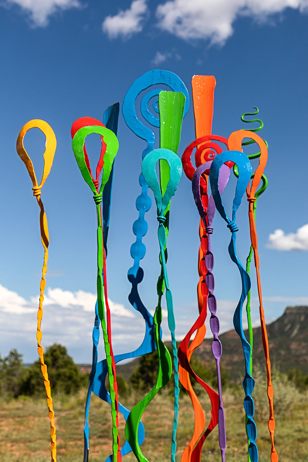 A group of different colored outdoor sculptures against a blue sky. Each sculpture has a slender stem with an abstract shape at the top.