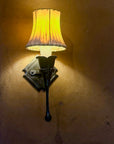 An old world style iron wall sconce with a hand forged calla lily holding a candle light bulb with a sheepskin shade mounted to an adobe wall.