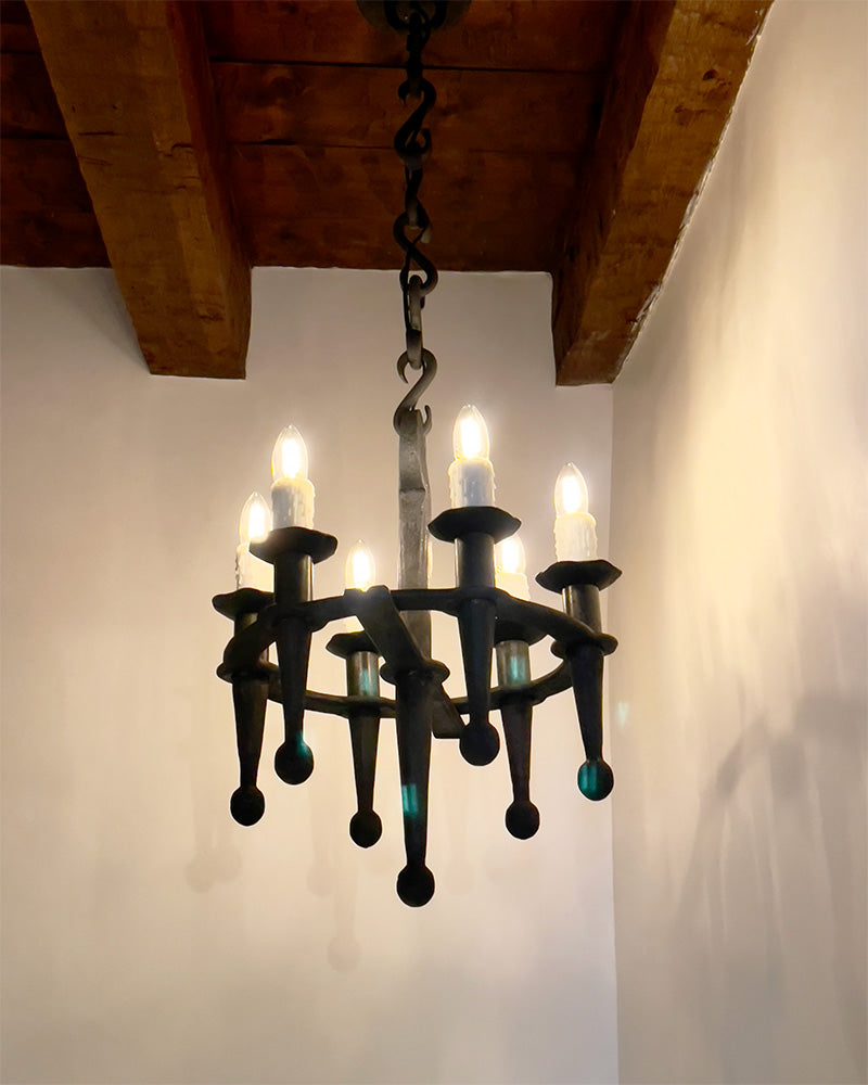 Wrought Iron Small Rustic Fleur Chandelier hanging from a ceiling. Six hand forged torches hold candle style light bulbs in a circle. It hangs from a handmade wrought iron chain made of s-hooks.