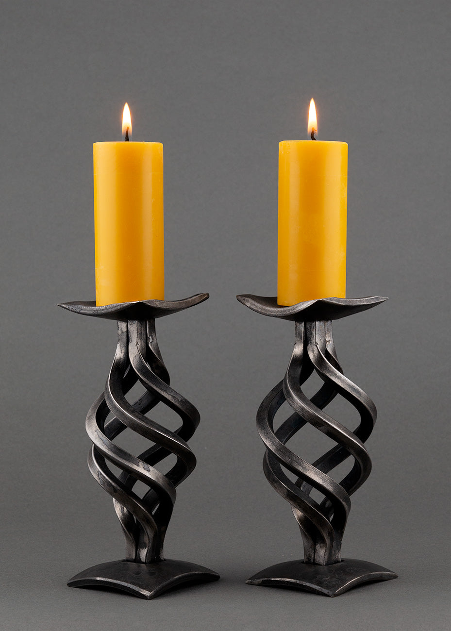 Two Swirl Wrought Iron Candle Holders with a square base. Four swirling pieces of forged steel make the stems. Shown with two burning beeswax pillar candles.