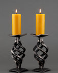 Two Swirl Wrought Iron Candle Holders with a square base. Four swirling pieces of forged steel make the stems. Shown with two burning beeswax pillar candles.
