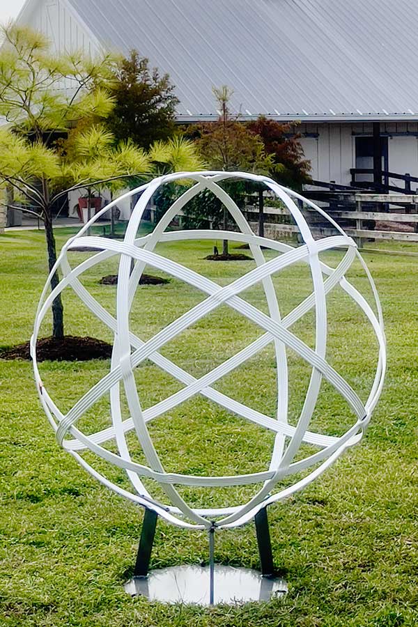 A multistranded white powder-coated orb sculpture on a three-legged stand, displayed on a lawn in front of a home.
