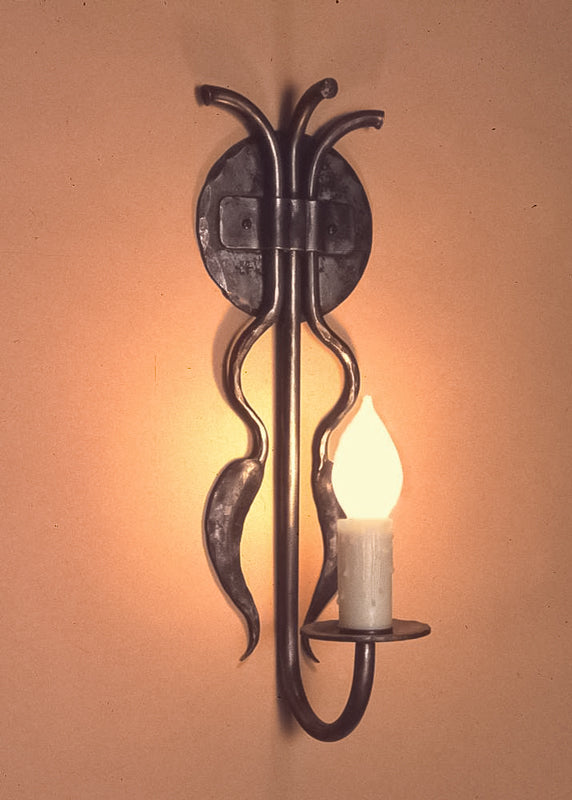 Hand-forged wrought iron candle wall sconce with two chili-shaped accents hanging from the backplate, featuring a candle-shaped bulb, in a rustic Southwestern style.