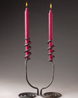Two artistic hand forged wrought iron candlestick holders with a swirling base that reaches upwards and wraps around the candles.