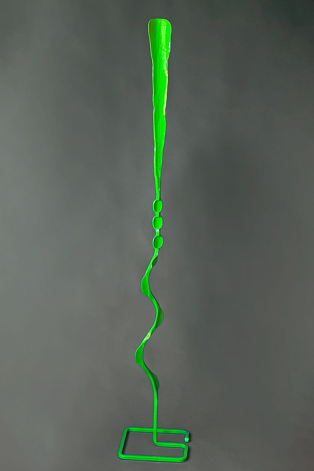 A vivid green, abstract-shaped steel sculpture with a long pole and a square free standing base, designed for indoor or outdoor decoration.