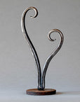 Opening Heart Sculpture made from hand forged steel. This small sculpture has a round steel base holding a heart shape sculpture that remains open at the top with each side of the heart curling inwards.