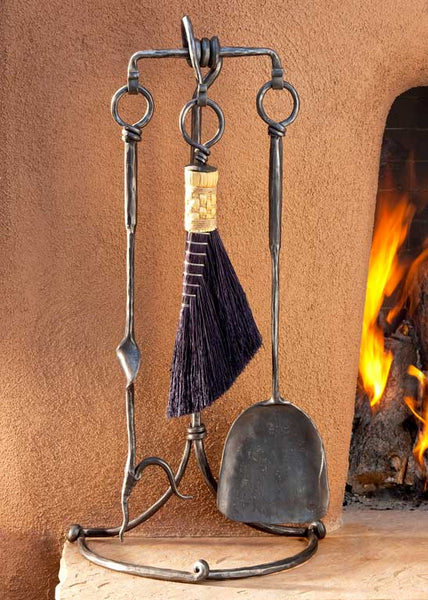 Heavy duty outdoor wrought iron fireplace tools with a broom, shovel and a poker standing next to a kiva fireplace.