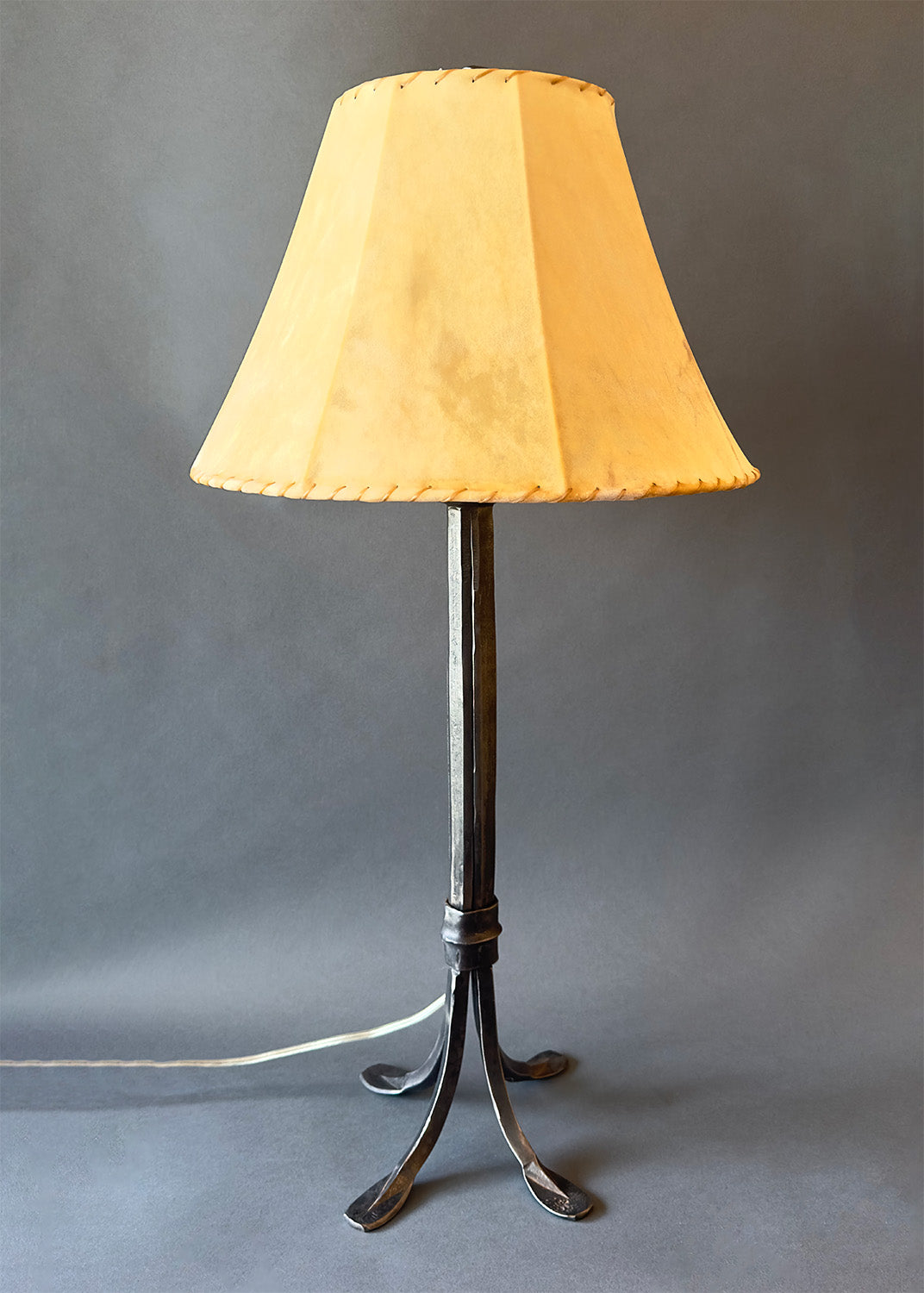 Hand-forged Collard Sheave wrought iron lamp featuring a tapered, black iron base with four elegantly curved feet. The lamp has a warm, tan lampshade with stitched edges, contrasting the dark metal and giving it a rustic yet refined appearance.