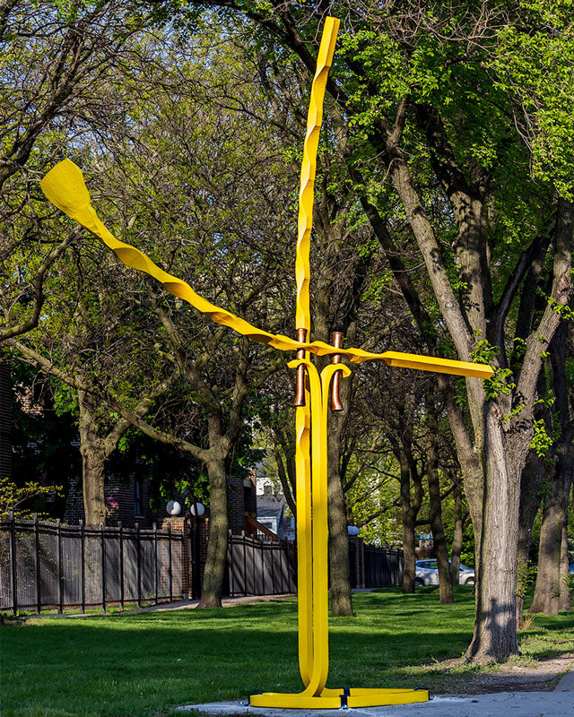 A bright yellow large abstract metal sculpture stands on a grassy park lawn surrounded by mature trees with green foliage.