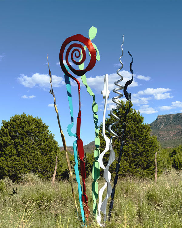 Abstract metal sculpture garden art piece with spiraling and wavy forms in red, turquoise, green, white, and black, set outdoors in a grassy field with trees and mountains under a blue sky.