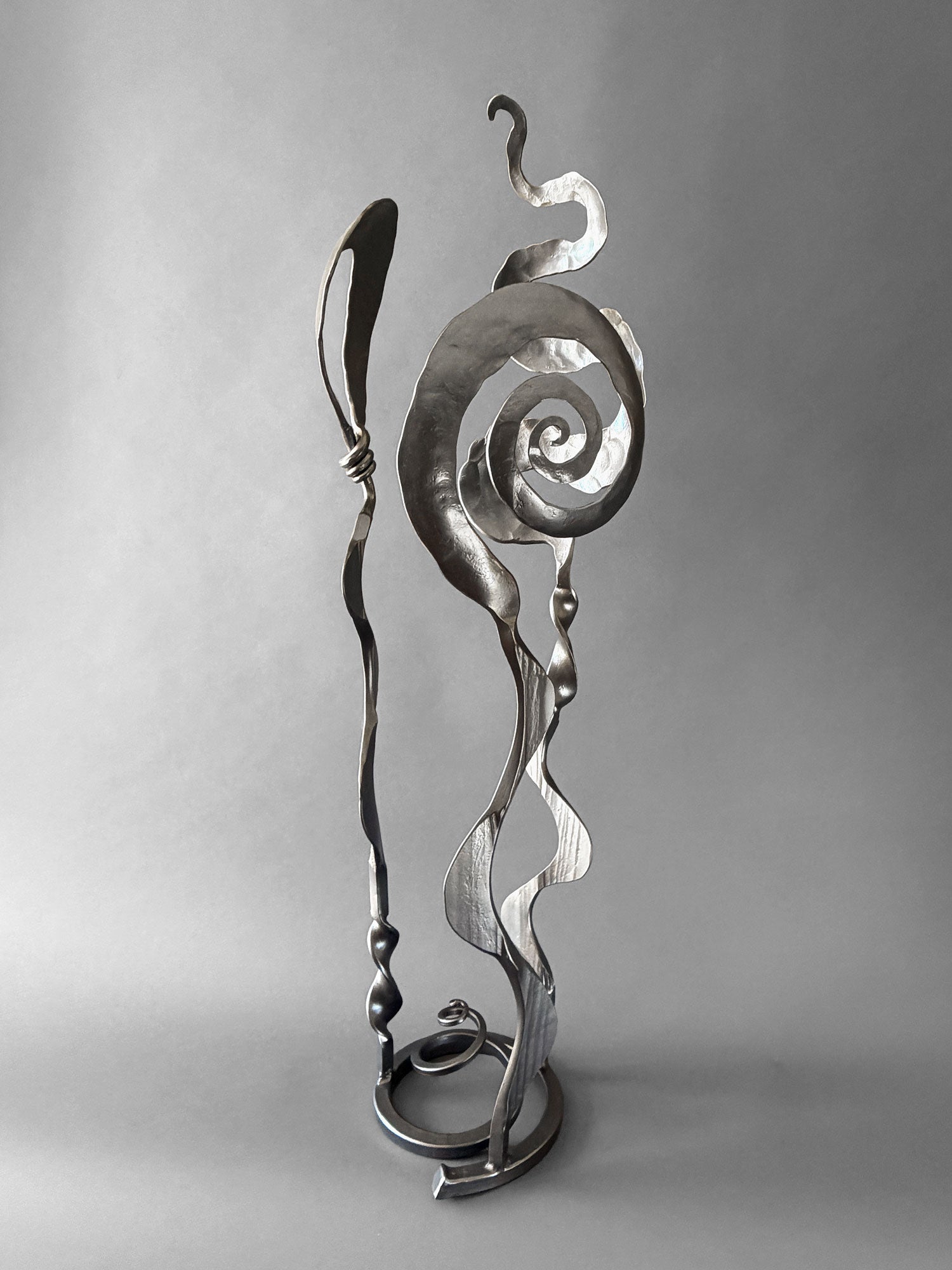 Abstract metallic sculpture with flowing curved and spiral forms on a circular base, photographed against a smooth gray studio background.