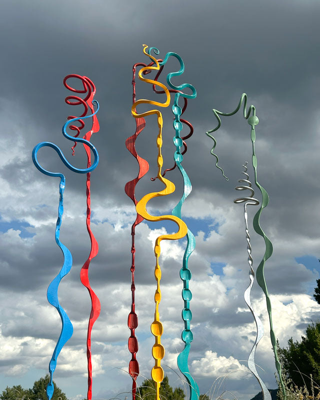 Seven tall, abstract metal ribbon sculptures in blue, red, copper, yellow, teal, silver and green, set outdoors against a dramatic cloudy sky.