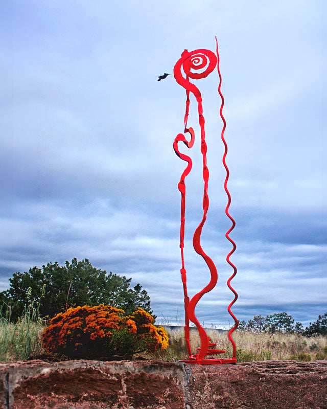 A tall, abstract red metal sculpture stands outdoors on a stone wall against a cloudy blue sky, with shrubs and trees in the background.
