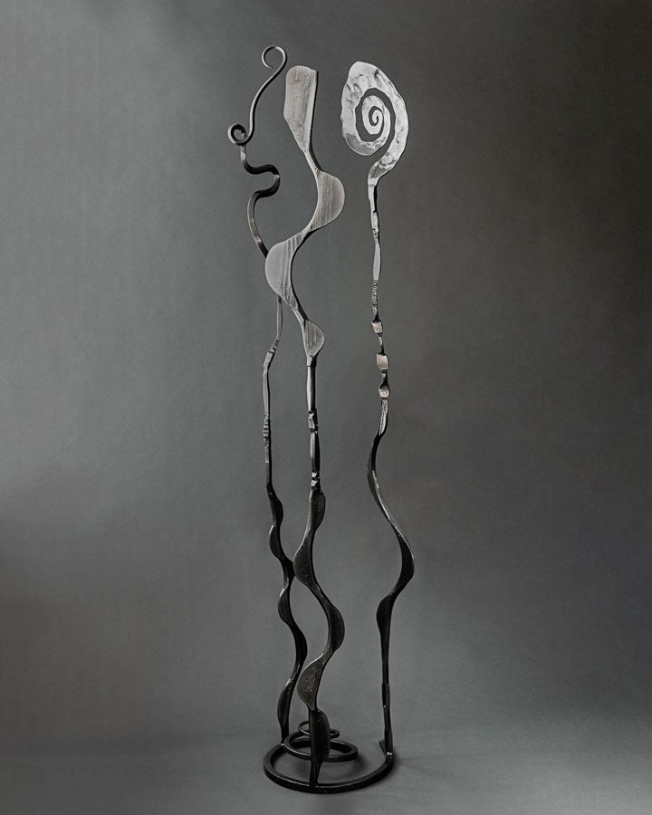 Abstract metal sculpture with three tall, wavy stems ending in spiral and looped and wavy 
forms, mounted on a round base against a dark gray studio background.