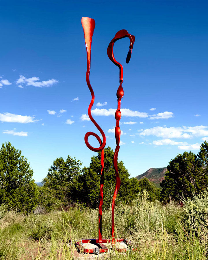 A tall abstract red metal sculpture stands outdoors in a grassy field under a blue sky with clouds and trees in the background.