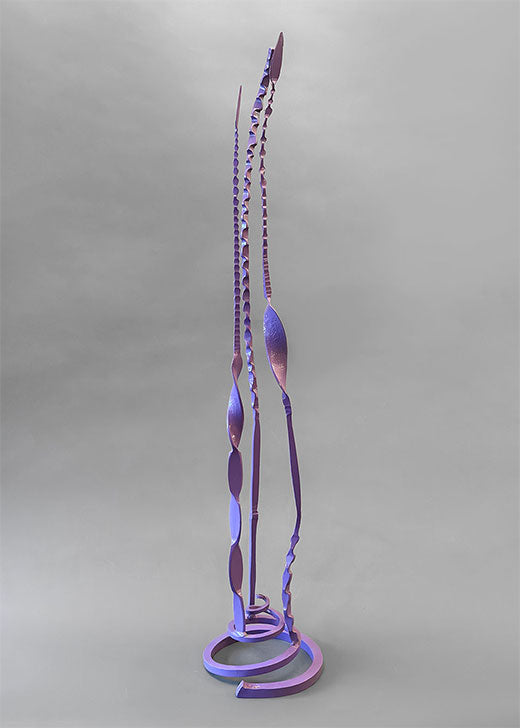 A tall abstract metal sculpture with a spiral base and elongated leaf- or blade-like forms, shown against a plain light gray studio background.