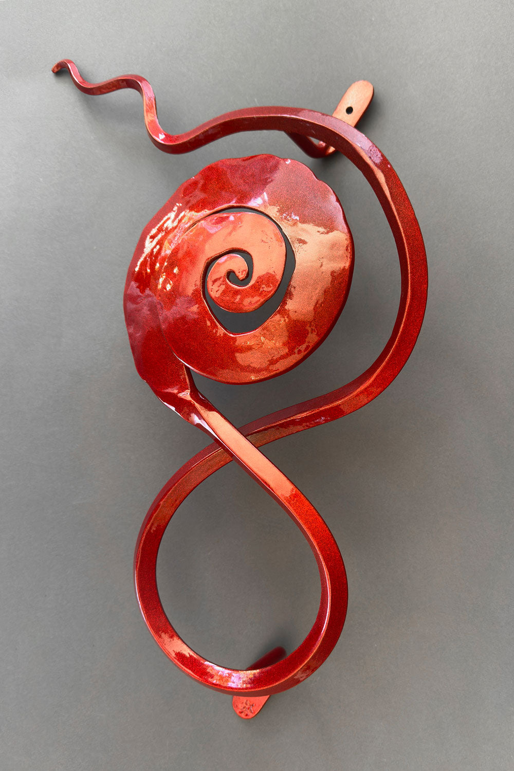 Hand-forged steel wall sculpture with a spiral rose design in a glossy copper-red finish, featuring flowing curves and mounted against a neutral gray background.