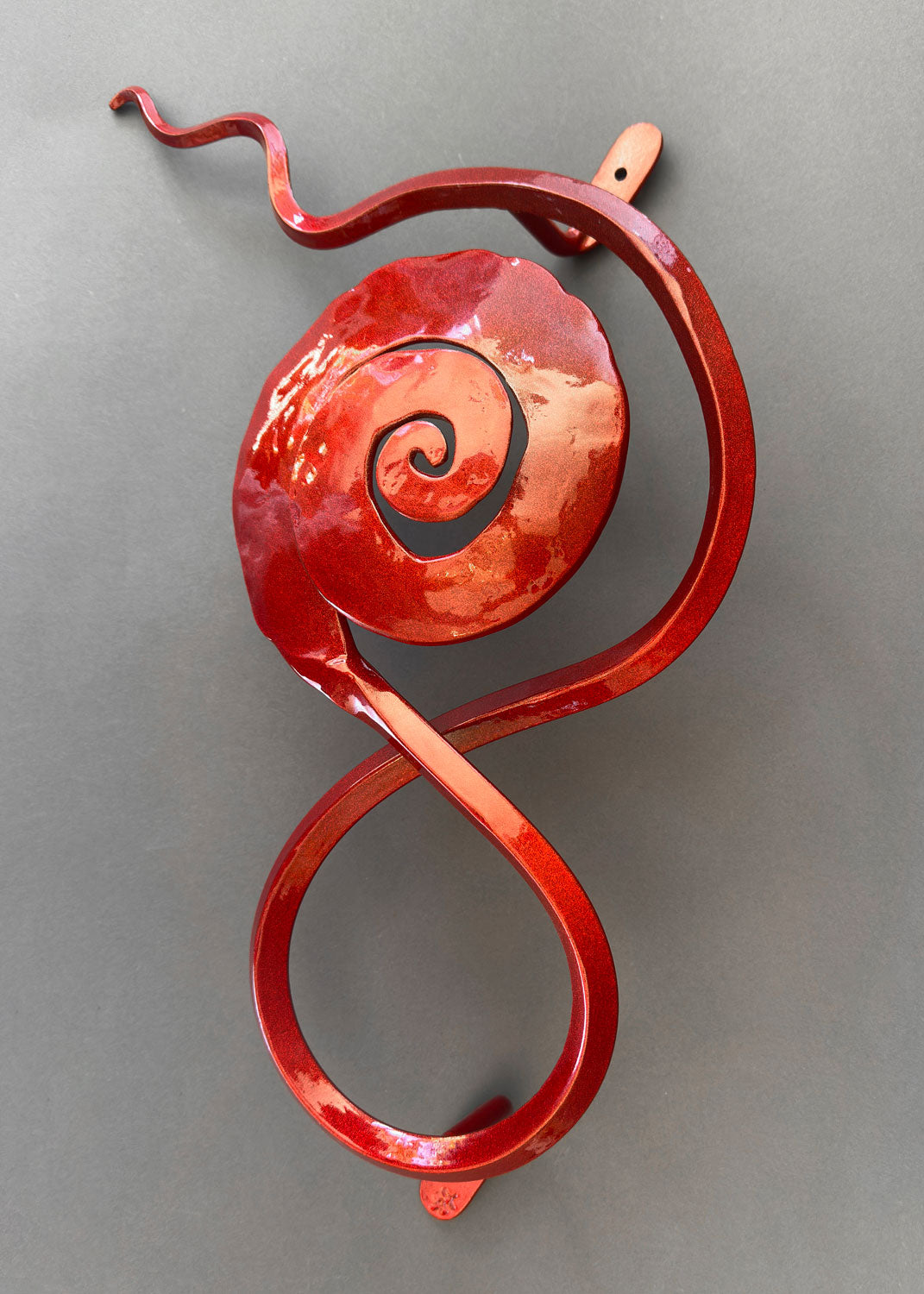 Hand-forged red metal wall sculpture with a spiral rose design in a glossy copper-red finish, featuring flowing curves and mounted against a neutral gray background.
