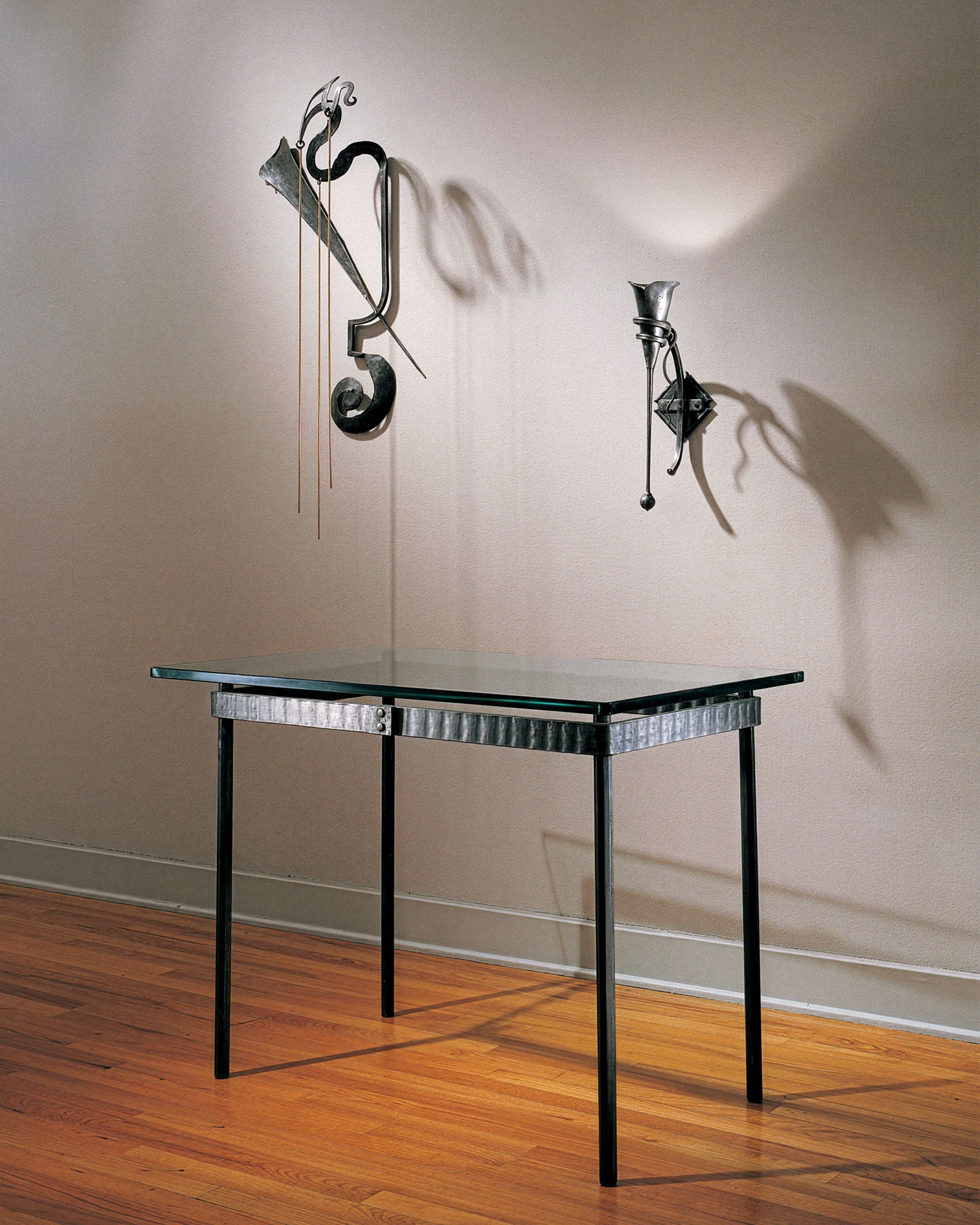 Modern glass top wrought iron writing table with decorative iron wall sconce and steel sculpture in a minimalistic setting. Hand forged ironworks by Christopher Thomson Ironworks.