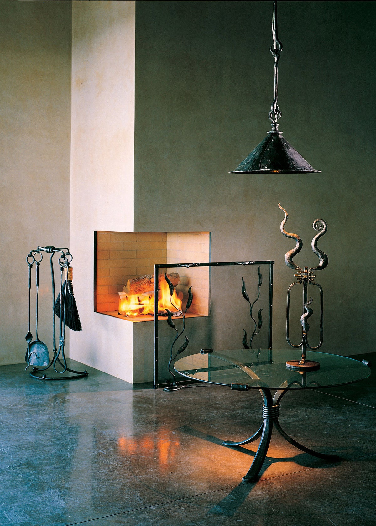 A modern fireplace with a lit fire, set in a minimalist interior with a standing fireplace tool set, glass-top table, hanging pendant light, and decorative metal sculptures, all hand forged by blacksmith, Christopher Thomson Ironworks.