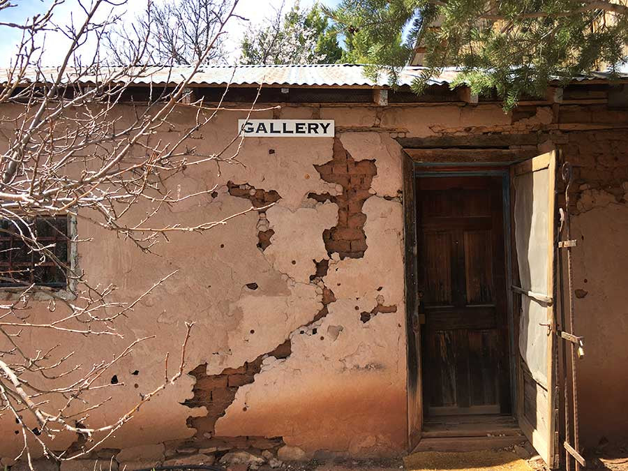 Weathered adobe wall with a rustic wooden door marking the entrance to a blacksmith gallery, sign reading 'GALLERY' above.