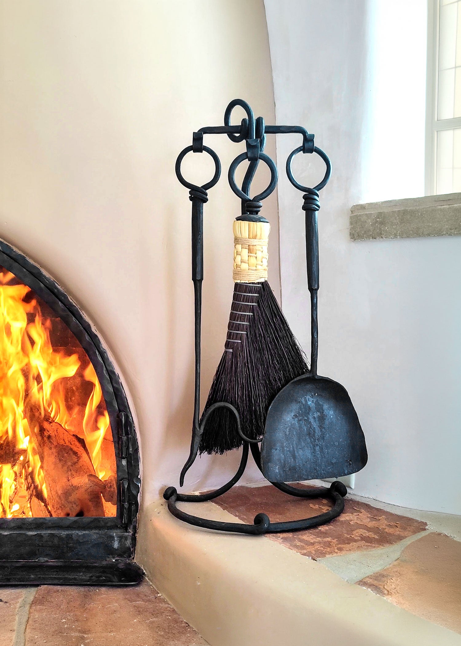A small rustic fireplace tool set made of hand forged steel, standing next to a kiva fireplace.