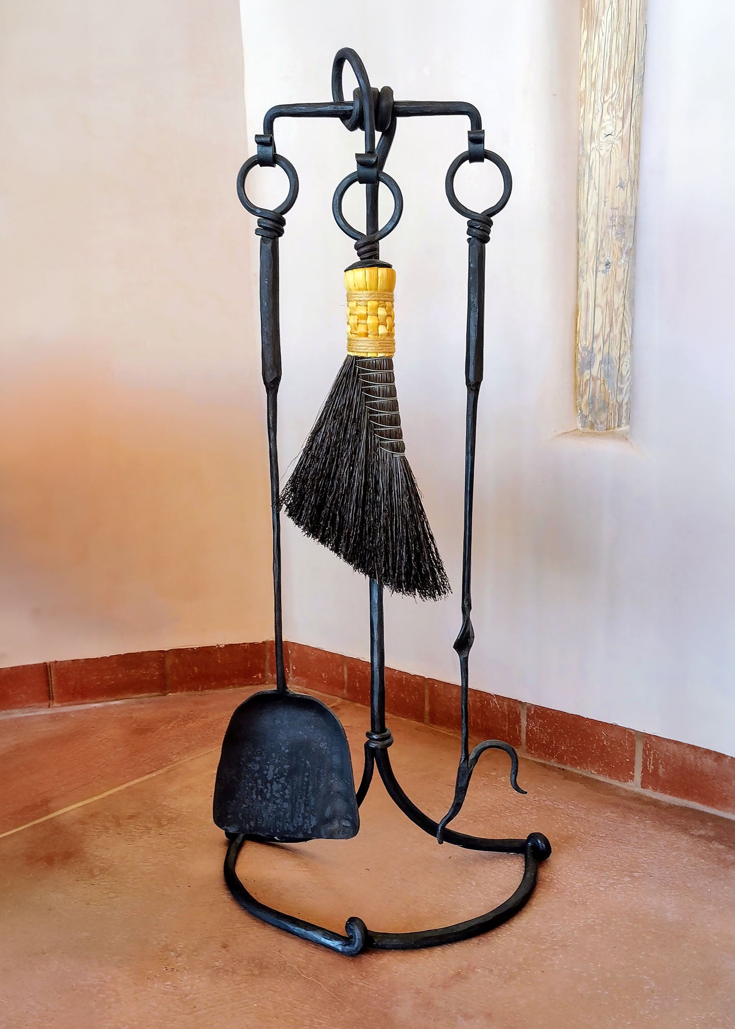 A rustic black iron fireplace tool set with a stand, including a shovel, poker, and broom displayed in a corner with terracotta tile flooring and cream walls.