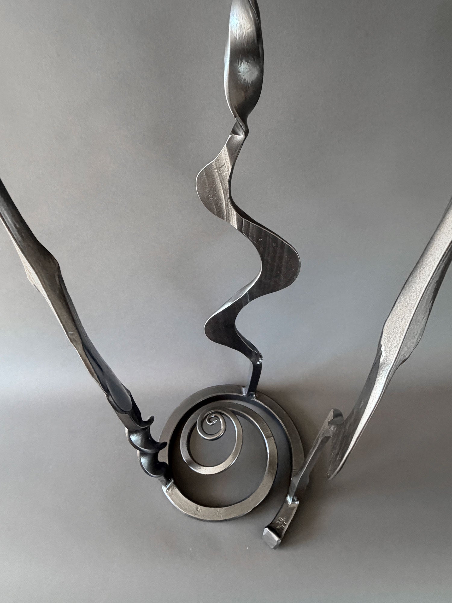 Close-up view of the lower portion of a metallic steel Baby Bloom sculpture showing the forged spiral base and three sculptural stems.