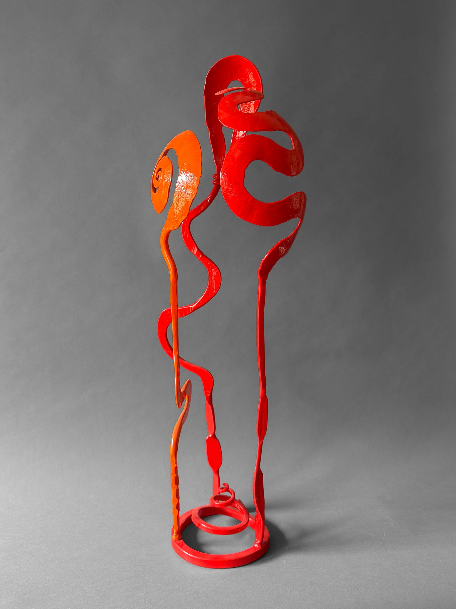 Orange and red hand forged steel Baby Bloom sculpture, front view, with large orange Rose spiral, red Meander and red Loop forms on circular base, 33 inches tall