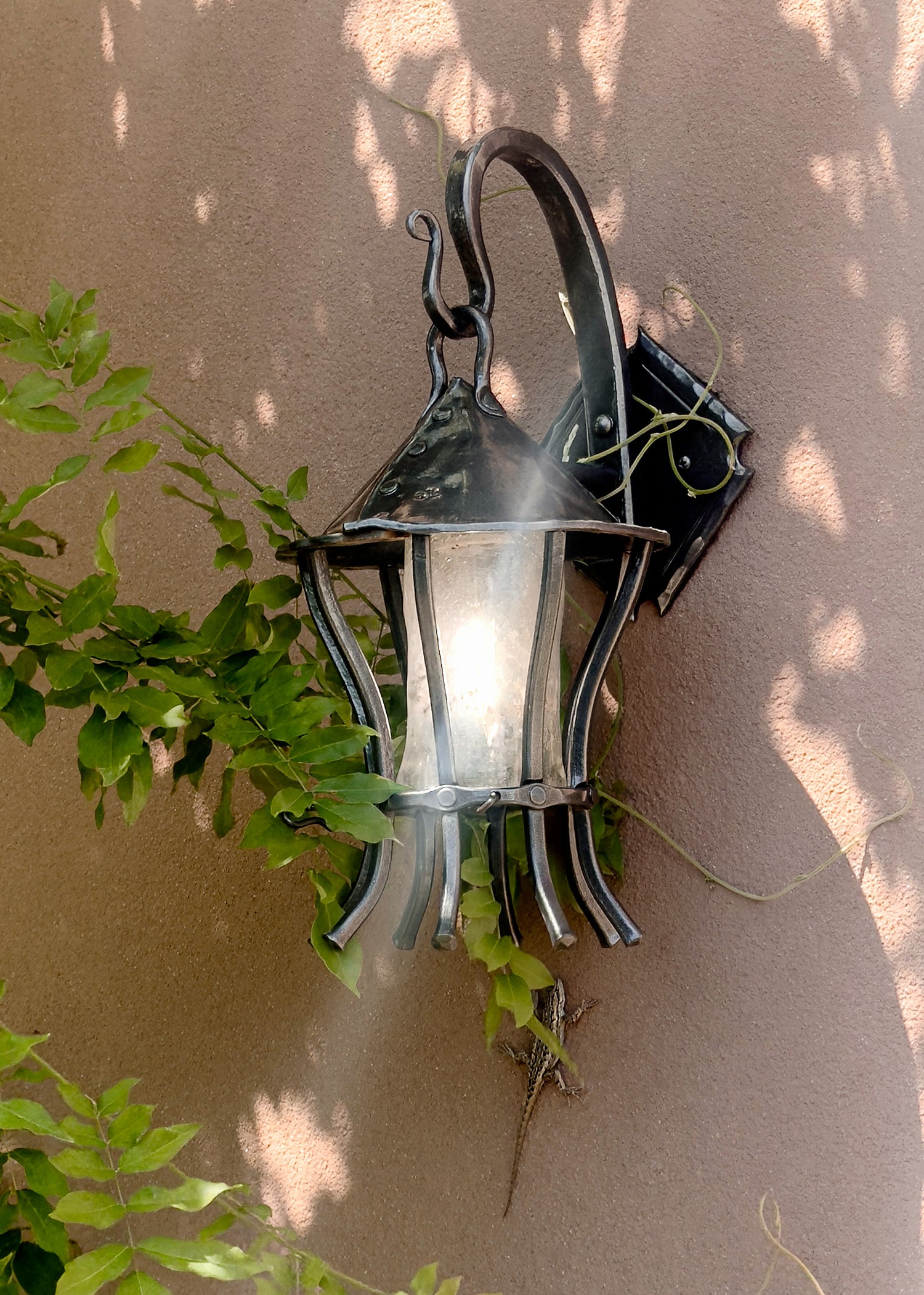 Hand forged wall lantern with hand blown glass mounted on an adobe exterior wall, surrounded by climbing greenery