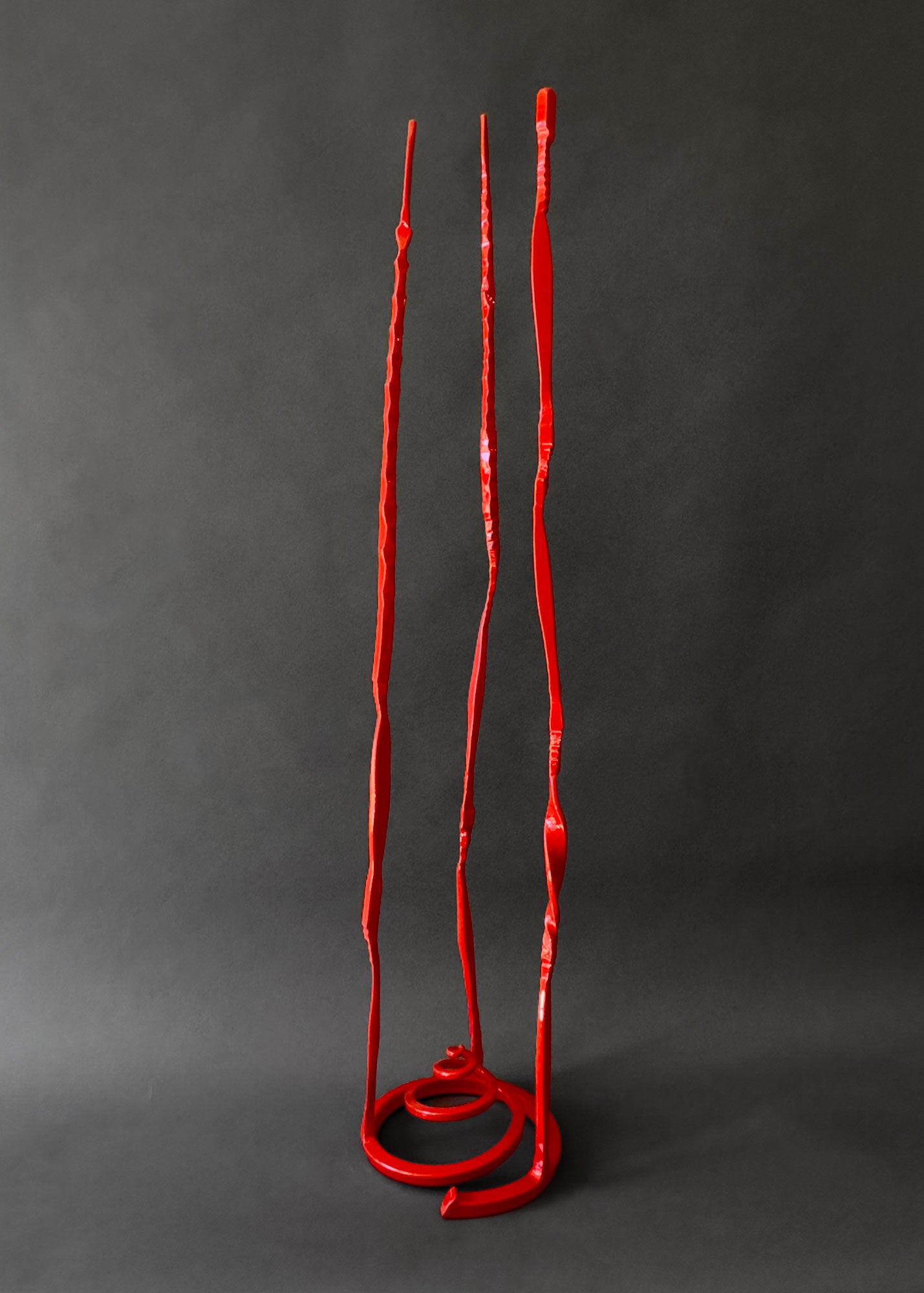 Small red steel sculpture with a spiral base and three slender vertical pieces welded to the base.