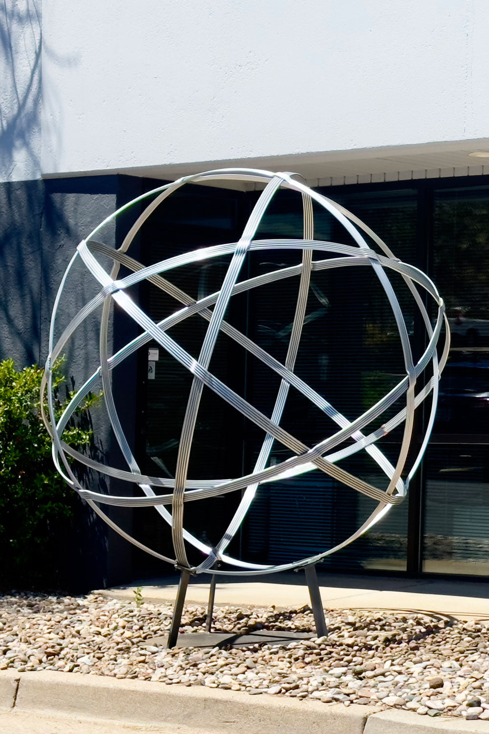 Large 76 inch spherical chrome sculpture, Chinlone Garden Orb, resting on a stand in front of a public building.