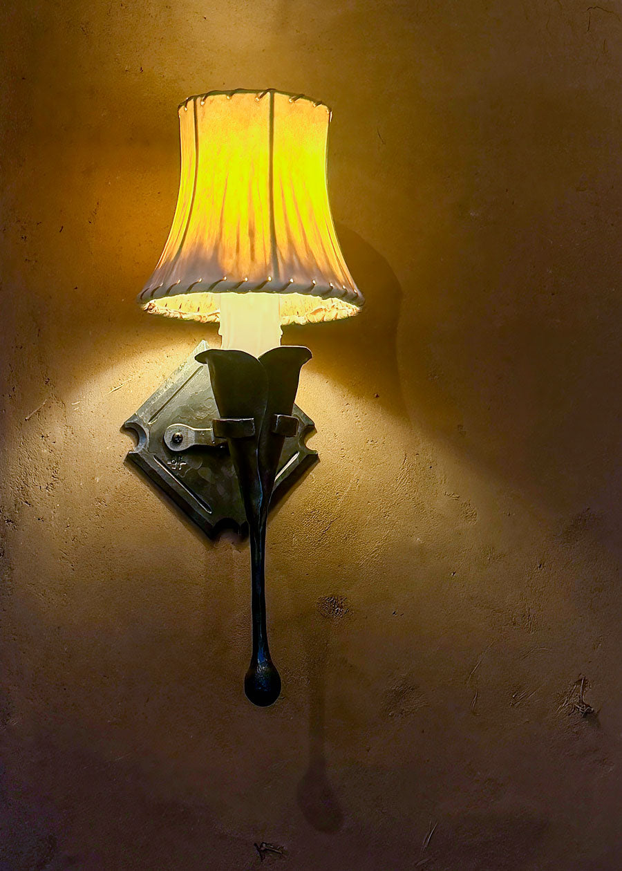 An old world style iron wall sconce with a hand forged calla lily holding a candle light bulb with a sheepskin shade mounted to an adobe wall.