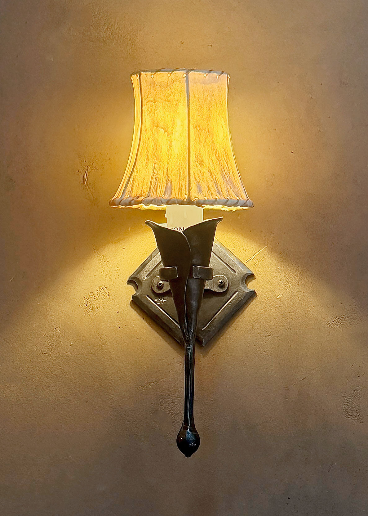 A small wrought iron calla lily wall sconce with a sheepskin shade mounted to an adobe wall with a diamond shaped backplate.