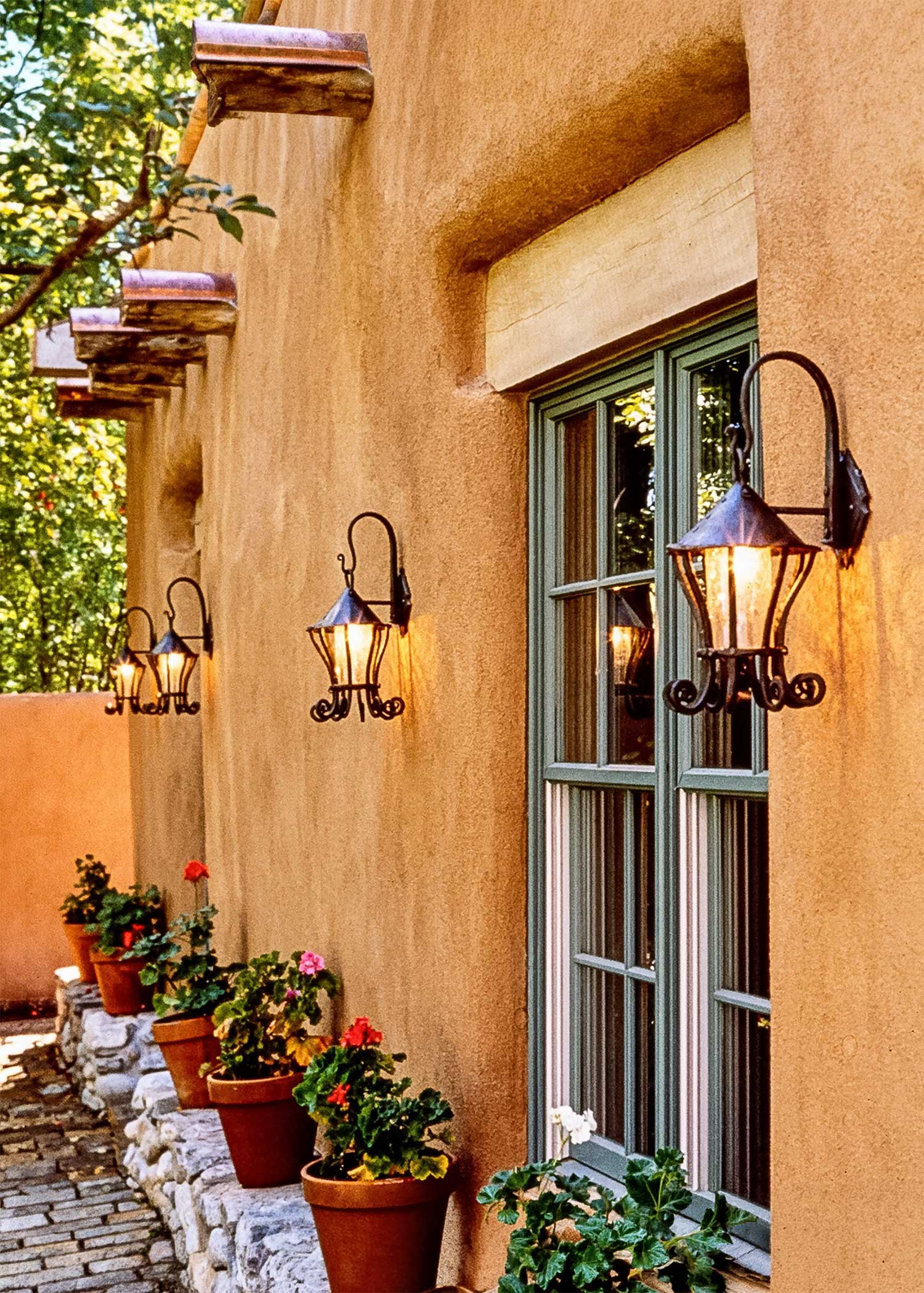 Small hand forged wrought iron hanging wall lanterns installed on a New Mexico adobe courtyard wall, glowing warm crackle glass light at dusk