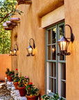 Small hand forged wrought iron hanging wall lanterns installed on a New Mexico adobe courtyard wall, glowing warm crackle glass light at dusk