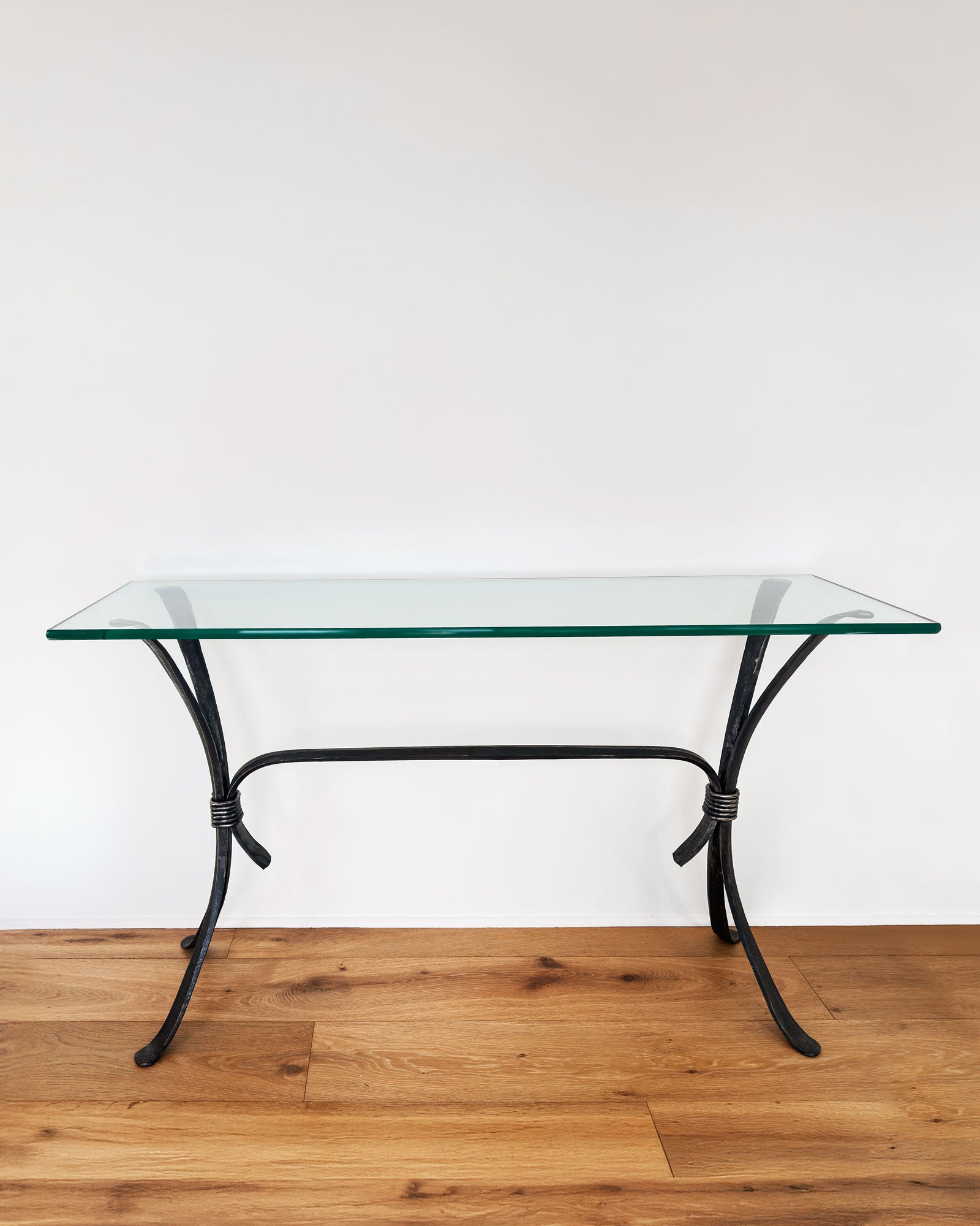 Wrought Iron Sofa Table with a glass top and a forged steel base on a wood floor against a white wall. 