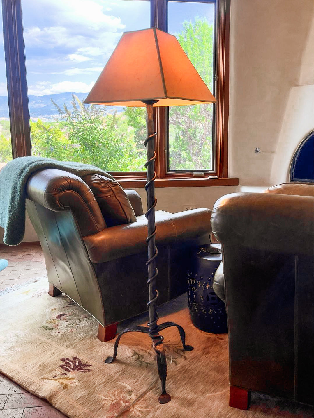A Wrought Iron Vine Lamp in a Southwestern style living room with a leather chair, couch, kiva fireplace and view of trees outside a window.