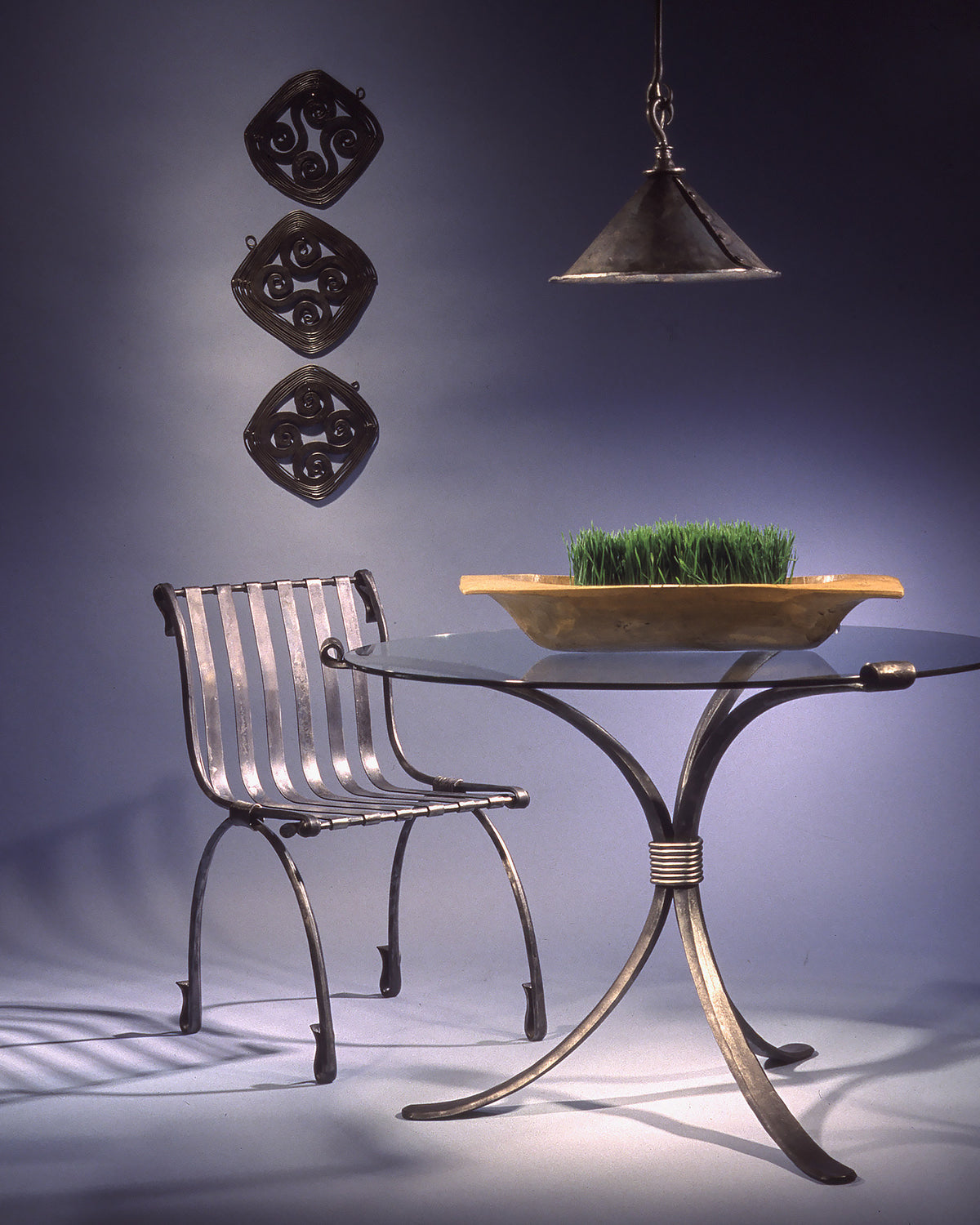 Round wrought iron dining table with a glass top on curved legs bound by a forged collar, beside an iron chair
